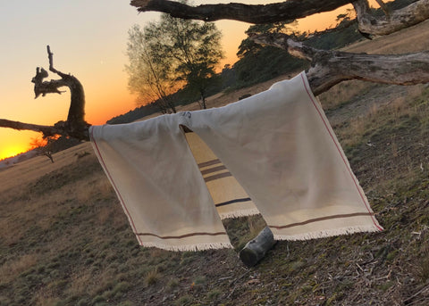 PICTURE OF PONCO LORIGA, MADE IN BEAUTIFUL WOOLWHITE. THE PONCHO IS MADE IN 50% BIO COTTON, 50% BIO WOOL. THE PONCHO HAS A RED LISTING, AND A BEAUTIFUL STAMP EMBROIDERED IN RED ON THE LEFT FRONT PANEL (FOR THE WEARER). STRIPED DETAILS IN BEIGE AND (DARK) GREY.