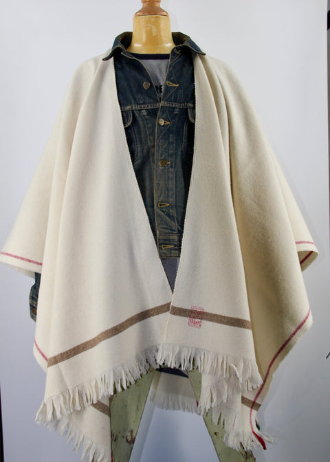 PICTURE OF PONCO LORIGA, MADE IN BEAUTIFUL WOOLWHITE. THE PONCHO IS MADE IN 50% BIO COTTON, 50% BIO WOOL. THE PONCHO HAS A RED LISTING, AND A BEAUTIFUL STAMP EMBROIDERED IN RED ON THE LEFT FRONT PANEL (FOR THE WEARER). STRIPED DETAILS IN BEIGE AND DARK GREY.